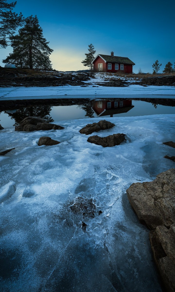 A cabin sits among trees at the edge of a still mountain lake in winter.