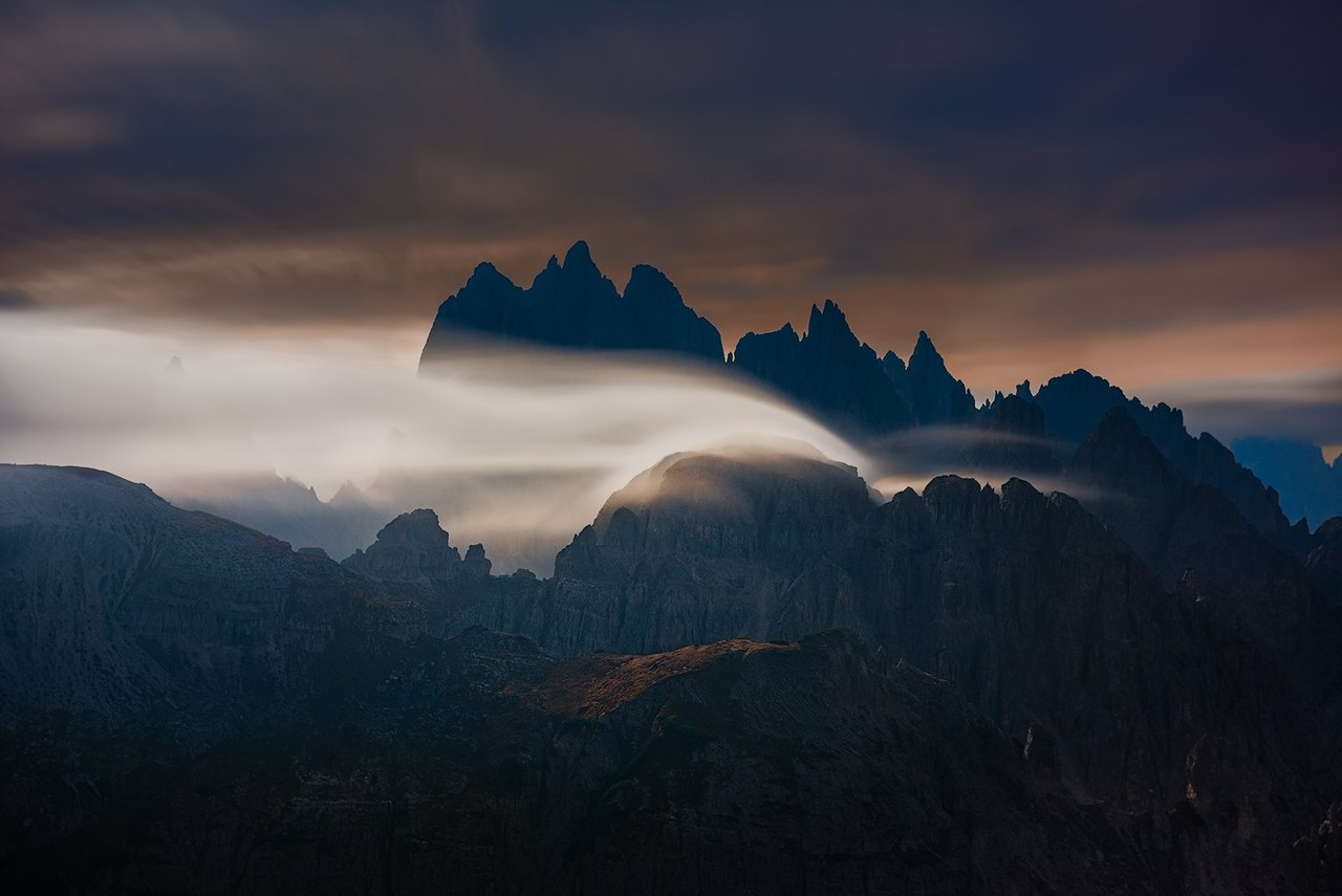 Jagged mountain peaks rise through a band of cloud beneath a muted evening sky.