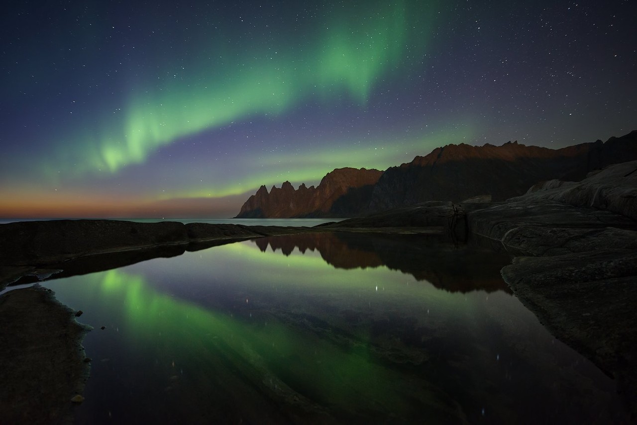 The northern lights glow above dark mountain silhouettes reflected in still water.