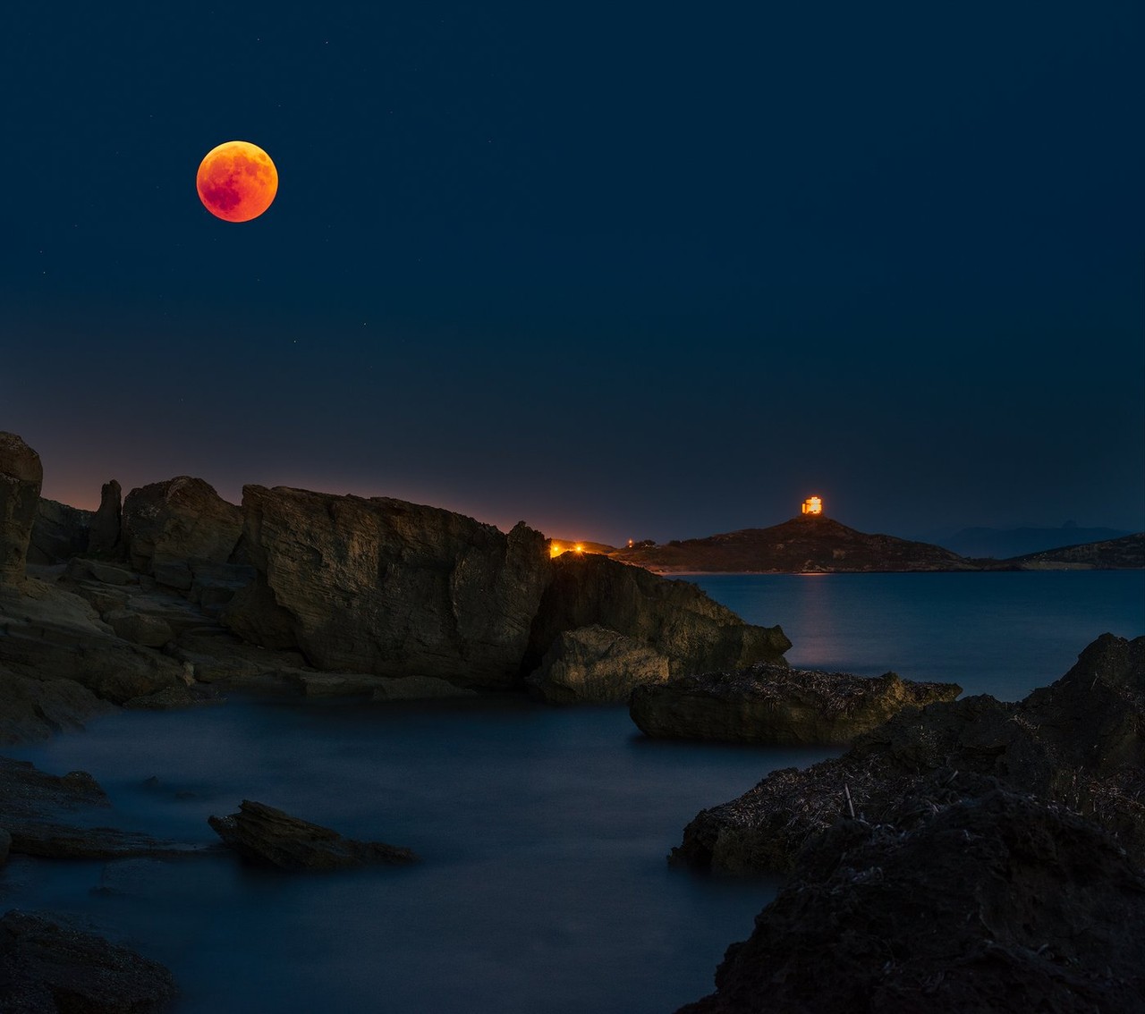 A red moon hangs above a dark Sardinian shoreline and sea.