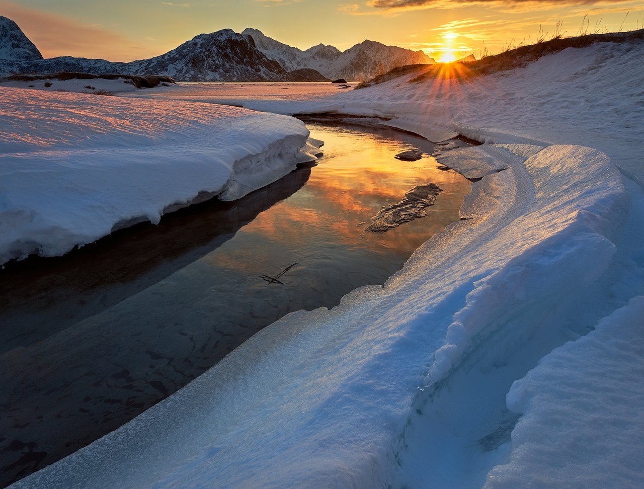A low winter sun reflects across a narrow inlet between snow-covered banks.