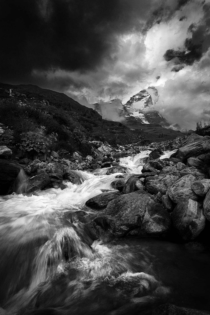 A rocky mountain stream leads towards the Matterhorn beneath dramatic storm clouds.