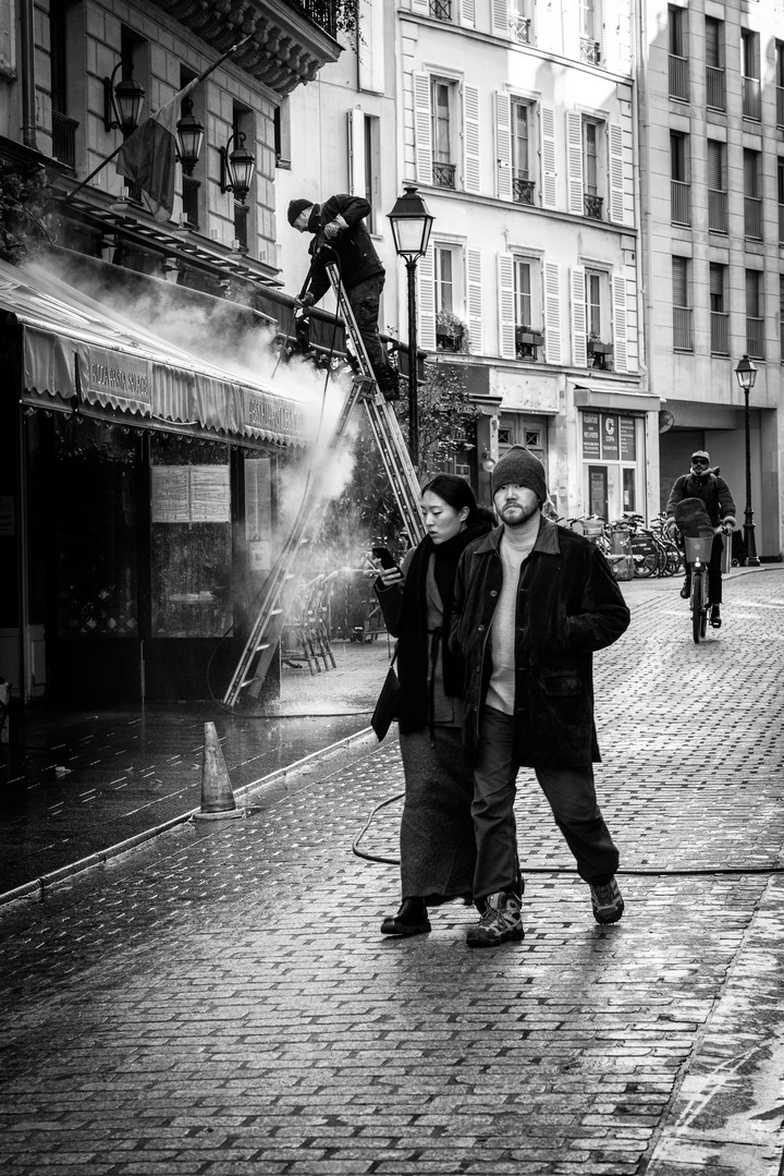 A worker stands on a ladder above a café awning as pedestrians cross a cobbled Paris street and a cyclist approaches.