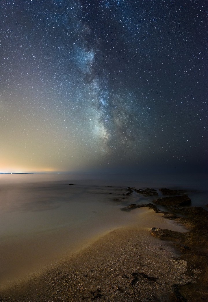The Milky Way rises above a dark rocky shoreline and the sea.