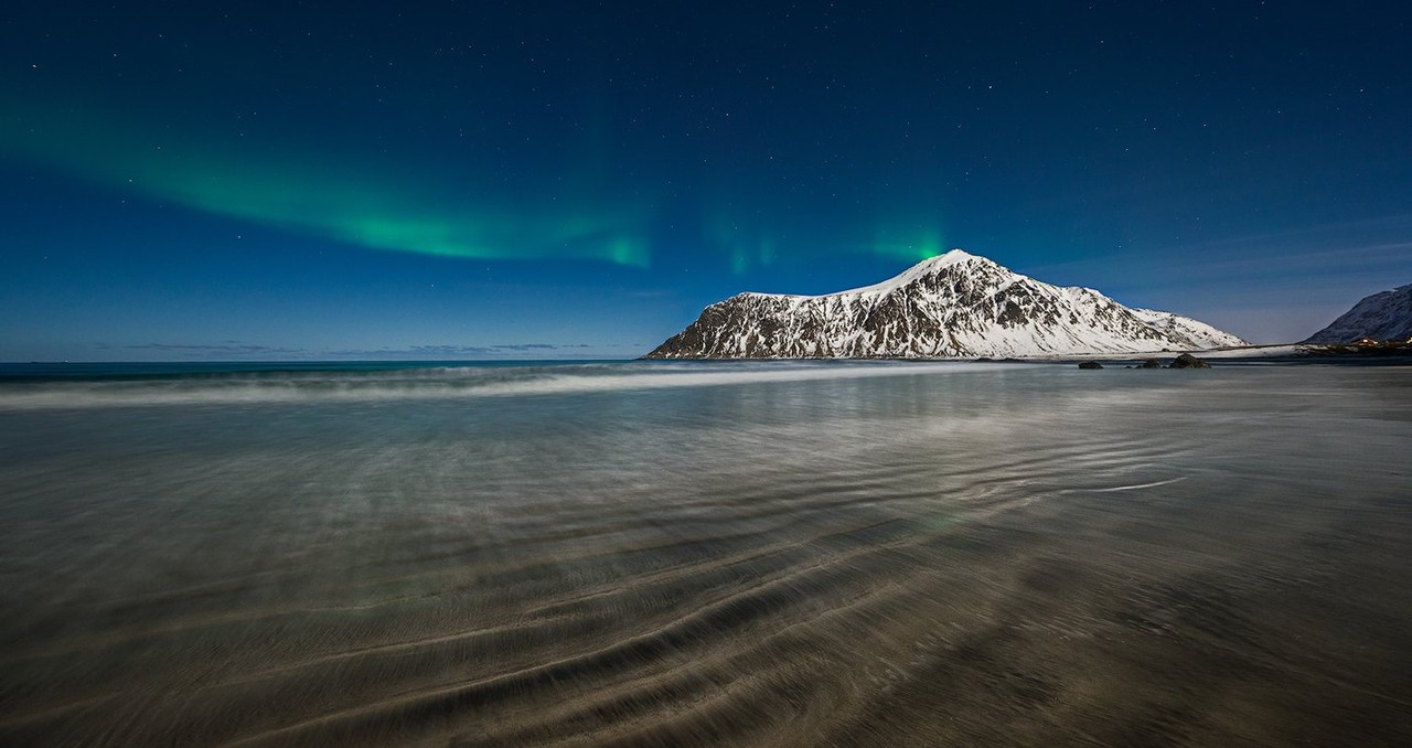 The northern lights glow over a mountain and its reflection on the shore.