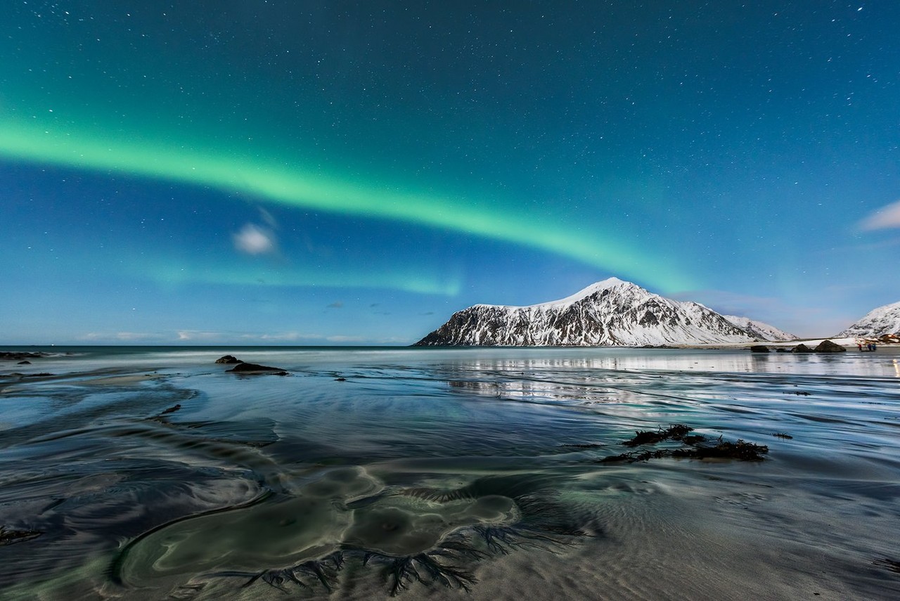 Green aurora stretches above a snowy mountain reflected in shallow water.